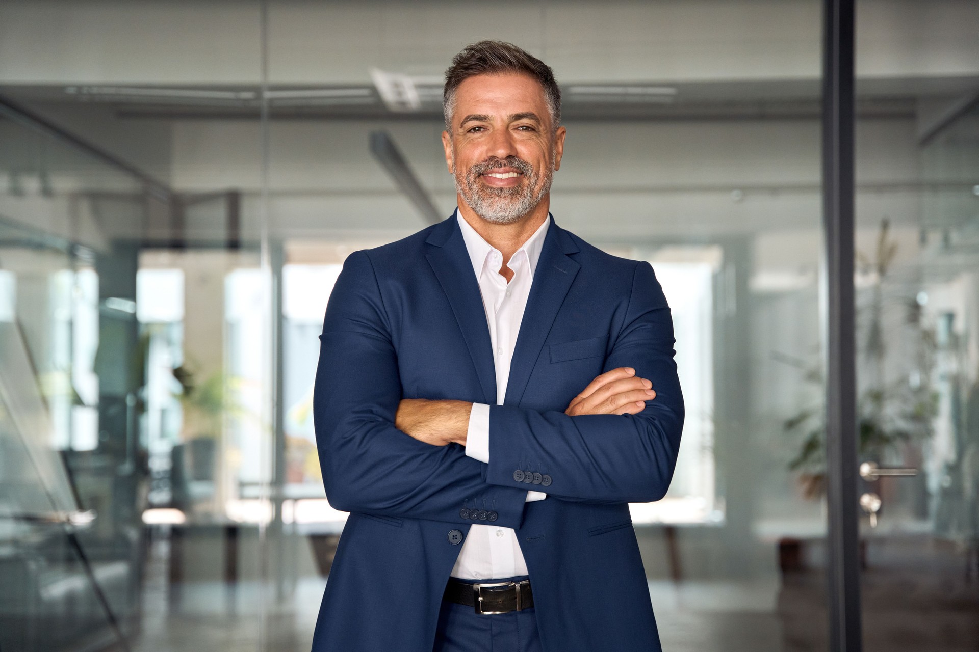 Handsome hispanic senior business man with crossed arms smiling at camera. Indian or latin confident mature good looking middle age leader male businessman on blur office background with copy space.